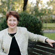 Suzanna Sheed in boucle blazer and Black dress sits on a wooden park bench smiling at the camera.