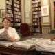 Suzanna Sheed MP, dressed in black business dress and white blazer site at a library table surrounded by papers.