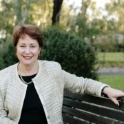 Suzanna Sheed pictured from the waist up sitting on park bench. She wears a black dress and cream boucle jacket and has one arm resting on the park bench. Suzanna is smiling at the camera.