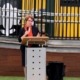 Suzanna in a dark suit and organe scarf address people at the opening of the children's centre. She is holding a microphone and standing behind a podium.